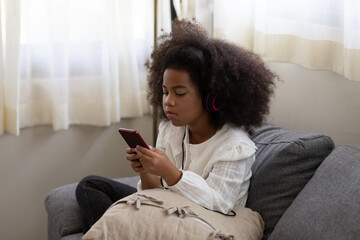African American little girl holding and looking at the mobile phone while sitting on sofa at home....