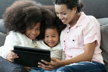 Happy afro family. Smiling African American mother with two little daughter looking at the digital tablet while lying on sofa at home. people, family, technology and education concept