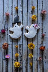 A lovely couple of sugar cookie ghosts wearing a hat and a flower on a weathered wooden background. There are a lot of colorful flowers on the background.