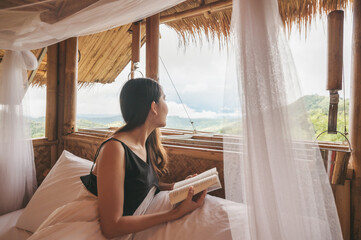 Young asian woman resting and reading a book on the bed in thatched hut among the mountain on vacation