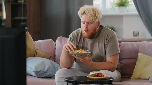 Young obese man sitting on sofa at home, eating fries with ketchup and watching TV