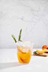 Recipe of cooking healthy summer peach juice - cocktail with ice, rosemary twig, sugar rim in glass, ingredients in elegant white interior on soft light wood table, vertical.