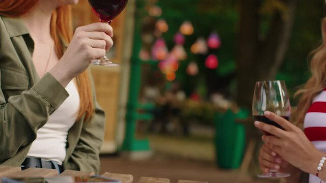 A close up shot of the wine glasses and a foxy red haired woman with an elegant blondie feeling cheerful while being outside at the park and sitting at a coffee table. 4k