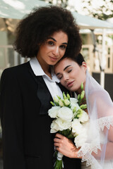 Young lesbian woman in veil holding bouquet near african american girlfriend in suit.