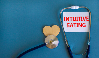 Intuitive eating symbol. White card with words Intuitive eating, beautiful blue background, wooden heart and stethoscope. Medical and intuitive eating concept. - obrazy, fototapety, plakaty
