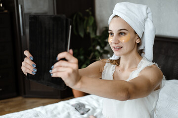 Beauty blogger, vlogger, influencer. Young Woman in pajamas, towel on her head and with cosmetic collagen patches under eyes recording video clip on tablet. Spa, skincare and wellness