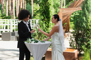 Cheerful interracial couple clinking with champagne during wedding outdoors.