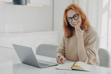 Positive ginger woman in casual clothes records audio message or smartphone while working on laptop