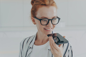 Redhead woman in transparent glasses speaks to virtual assistant holds mobile phone near mouth