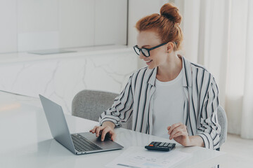 Young positive red-haired woman accounter working remotely on laptop and using calculator