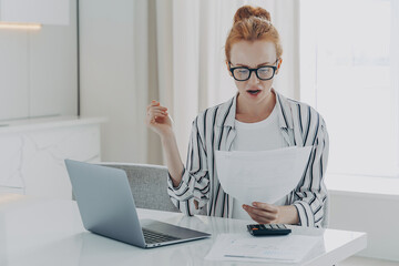 Savings finances concept. Shocked ginger woman focused at document studies bills reads banking paper