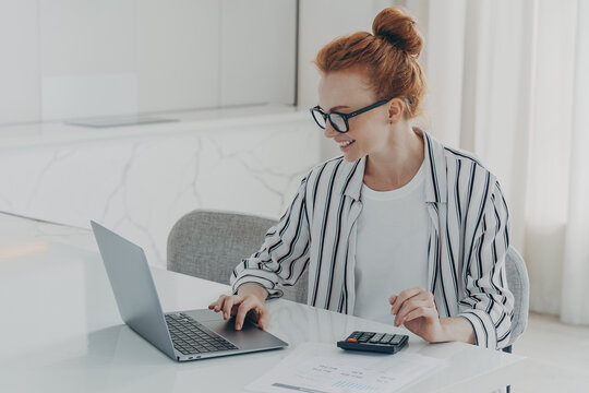 Young positive red-haired woman accounter working remotely on laptop and using calculator