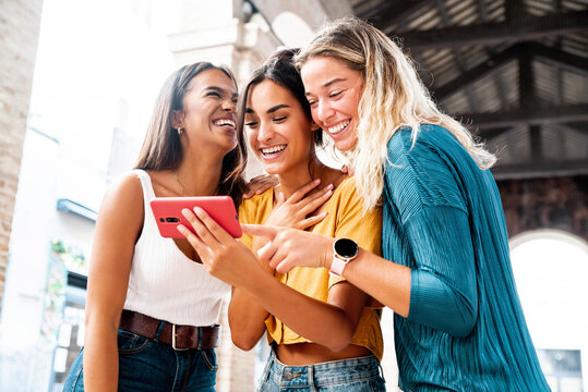 Three happy friends watching a smart phone mobile outdoors - Millennials women using cellphone on city street - Technology, social, friendship and youth concept