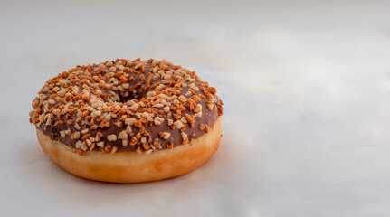 A chocolate donut with hazelnut sprinkles lies on a white surface. Fried dough confection, dessert food, sweet snack with chocolate glazing and nut crumbs. Delicious fresh donuts concept.