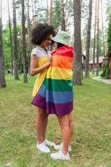 Positive african american woman hugging girlfriend in lgbt flag in park.