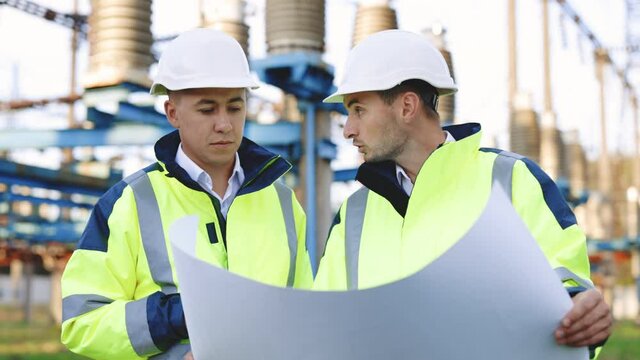 Two engineers are talking in front of electrical transmission lines, working on renewable energy development. Engineers in special clothing discuss a drawing on paper near high-voltage power line