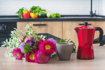 Bunch of multicolored asters, cup of coffee, red geyser coffee maker on wooden table in the kitchen