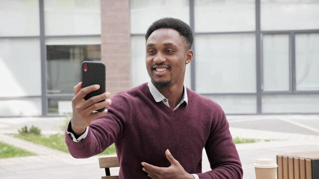 Afro American Businessman sitting in cafe near office center while having Video Call, Wearing Stylish Casual Clothes. Smiling and enjoying coversation. Business Building in the Background waving hand.