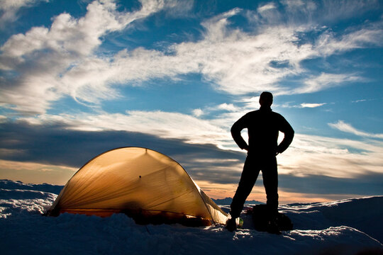 Silhouetted hiker standing next to backlit tent on mountain summit.