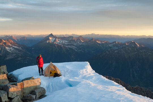 A climber stands by his tent, perched on the summit of a mountain.