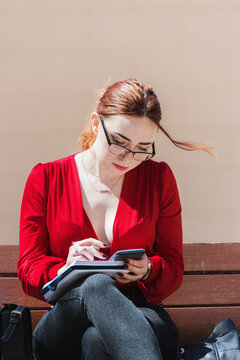 Young red-haired woman with glasses sitting on a bench while using a calculator and writing something in her notebook. Dressed in a red blouse.
