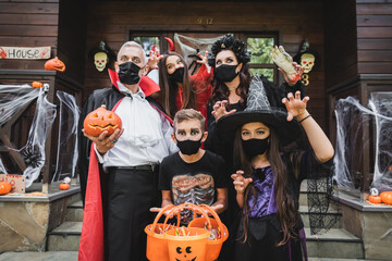 Fototapeta premium family in halloween costumes and black medical masks standing with buckets of sweets near house