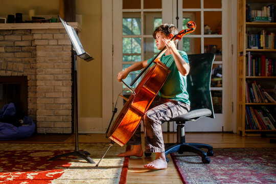 A barefoot boy practices cello in golden window light indoors