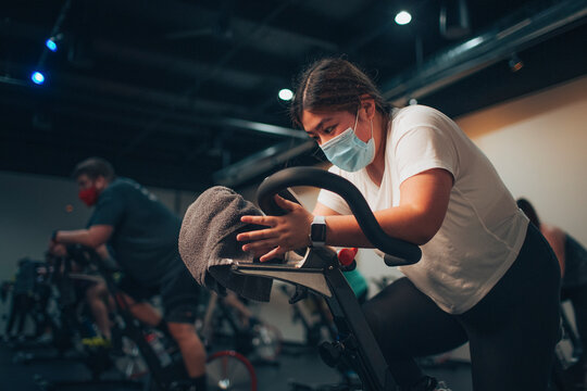 An asian woman pedals on a stationary bike in an indoor bike studio. - Powered by Adobe