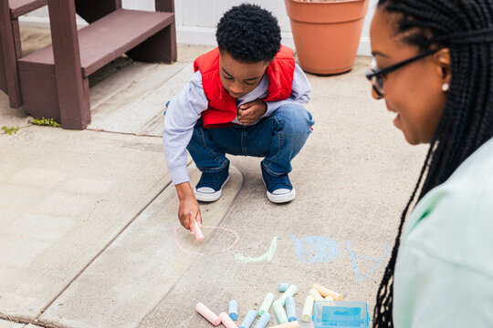 Boy using chalk to draw on concrete while mother smiles and helps
