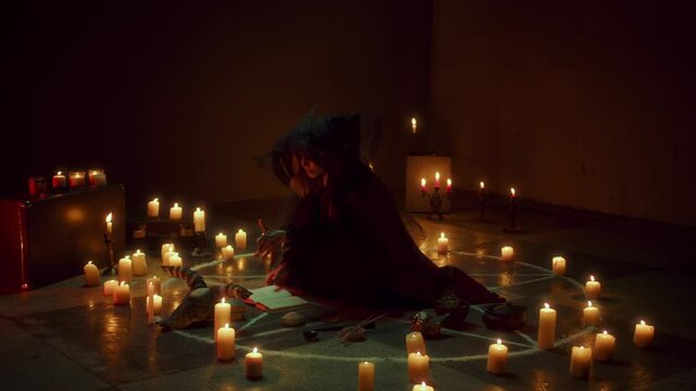 Witch woman sitting in pentagram circle and reading spell. Evil sorceress making rite and sacrifices at night, using black witchcraft, cow skull and candles on floor, shouting. Halloween time.