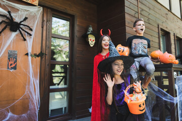 girl in witch hat waving hand near excited kids in spooky halloween costumes on porch with...