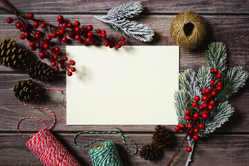 Blank white card surrounded by Christmas decoration on wooden background. Festive holiday greetings. Merry Christmas. Red berries, cones, twine, branches, ribbon. Empty space for text. Copy space.