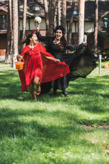 thrilled mom and daughter in halloween costumes running with skull, toy hand and bucket of candies