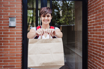 smiling rider woman wearing red backpack holding paperboard bag of food in city. Delivery service...