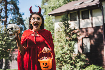 Obraz premium astonished child in demon costume screaming while holding skull and halloween bucket near blurred cottage