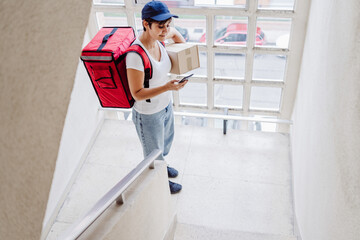 young rider woman wearing red backpack holding paperboard box in neighborhood stairs. Delivery service concept. Sustainable transport