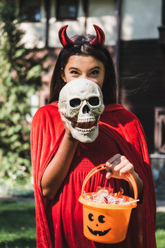 cheerful kid in devil halloween costume obscure face with spooky skull while holding bucket of sweets