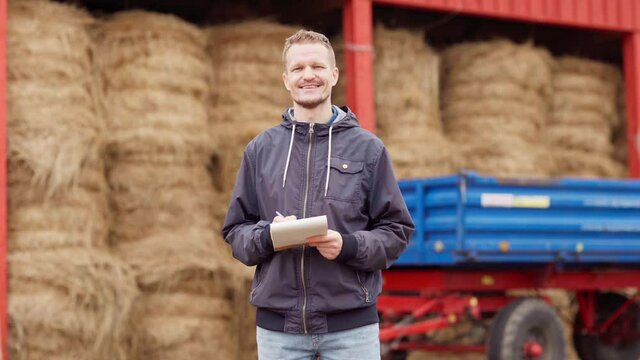 Tilt Down Portrait Of Middle Aged Farmer Or Agricultural Worker With Notepad And Pen Walking Towards Camera At Farm. Man Smiling Happily Standing Near Hay Bales Storage And Box Trailer