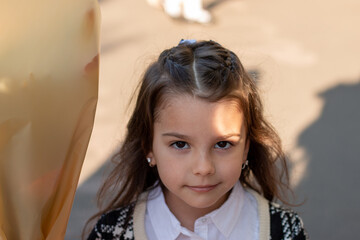 Fototapeta premium Smiling little girl with long dark hair in a white blouse and a plaid jacket holding a bouquet of flowers in her hand. First-grader's first bell at school