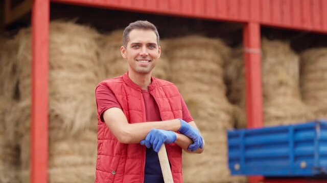 Tilt down portrait of young farmer or agricultural worker with pitchfork walking towards camera at farm. Man in workwear and gloves smiling near hay bales storage