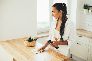 Young woman cleaning wooden table using spray and natural rag in a kitchen.