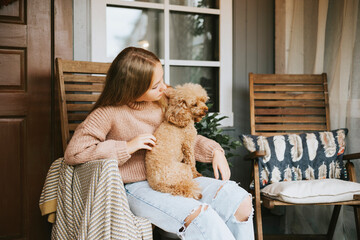 beautiful teenage girl in cozy warm knitted sweater sits and hugs with poodle dog on porch of backyard decorated with pumpkins and dry grass, cozy and comfortable autumn lifestyle concept