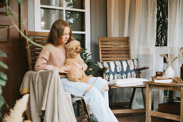 beautiful teenage girl in cozy warm knitted sweater sits and hugs with poodle dog on porch of...