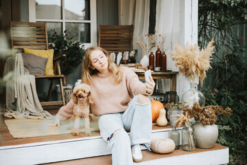 beautiful teenage girl in cozy warm knitted sweater sits and takes selfie on phone with poodle dog on porch of backyard decorated with pumpkins and dry grass, cozy and comfortable autumn lifestyle