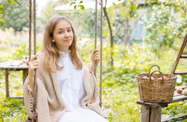 beautiful teenage girl in white shirt and knitted sweater sitting on swing in garden with basket full of ripe apples and apple in hand, concept picking fruit and harvest, cozy and comfortable autumn