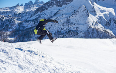 Chłopiec w stroju narciarskim wykonujący skok ze śnieżnej góry. Zimowy krajobraz górski, w tle skaliste, ośnieżone szczyty górskie. - obrazy, fototapety, plakaty