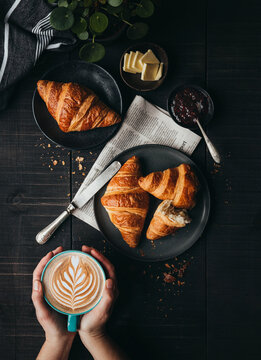 Hands holding latte beside croissants on black table from above.