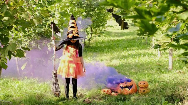 Puffs of purple smoke emanate from the carved eyes of the jack o lantern. A five-year-old girl in a witch costume with a broom stands among the maples next to the smoking purple color smoke. halloween