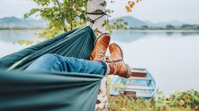 Young woman's leather fashion boots photo. Female swinging in a hammock between the birch trees on the mountain lake bank. Out-of-town Outdoor Recreation in Nature concept image.