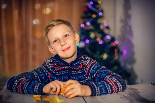 A boy with brown hair cleans a tangerine sitting near a decorated Christmas tree